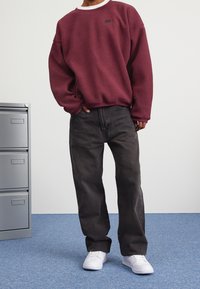 Sudadera de forro polar color granate con un logo negro, combinada con pantalones gris oscuro y zapatillas blancas. Diseño sencillo con un ajuste relajado.