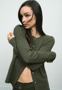 Olive zöld kardigán strukturált kötött mintával, kerek nyakkivágással és fa gombokkal, részben kigombolva az oldalon.