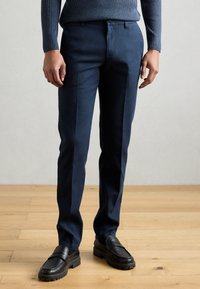 Marinblå skräddarsydda byxor med en slät textur, utrustade med en frontdragkedja och två sidofickor, kombinerade med svarta loafers.