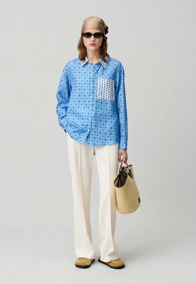 Camicia blu a fantasia con tasca bianca, pantaloni crema, infradito marroni e borsa intrecciata. Il modello indossa occhiali da sole e un leggero foulard.