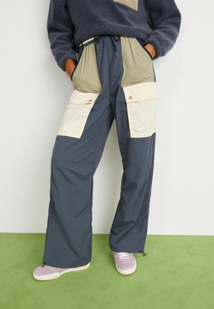 Personne portant un pantalon utilitaire ample à panneaux de couleurs avec de grandes poches avant et des poignets à cordon, assorti à un pull en tissu duveteux bleu marine et des baskets roses.