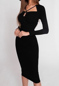 Donna che indossa un abito midi aderente nero a coste con maniche lunghe e dettagli incrociati sul collo, in piedi davanti a uno sfondo semplice.