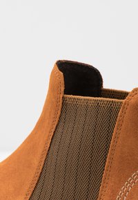 Zbliżenie na beżowy zamszowy but z czarną elastyczną boczną wstawką, która ma wzór jodełki i widoczne szwy.