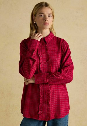 Red plaid button-up shirt featuring a ruffled front detail, long sleeves, and a classic collar, paired with blue jeans.