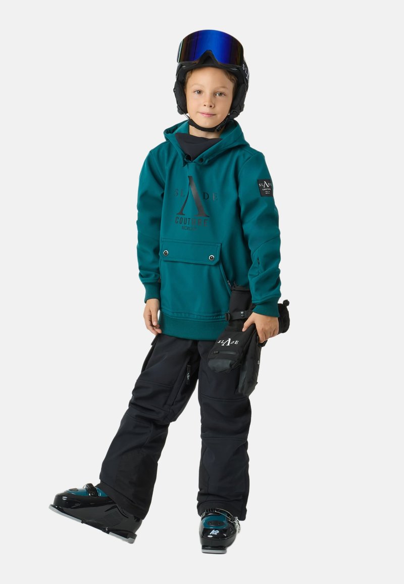 Felpa con cappuccio turchese con logo, due tasche frontali, pantaloni da sci neri e scarponi da sci neri. Indossa un casco con visiera a specchio, mani che tengono l'attrezzatura.