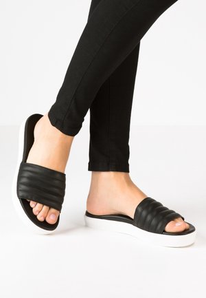 Voeten die zwarte doorgestikte instapsandalen met witte zolen en een zwarte broek dragen tegen een witte achtergrond.