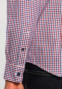 Červená, bílá a modrá kostkovaná košile s manžetou na knoflíky. Materiál je tkaná bavlna s hladkou texturou a kontrastními tmavými knoflíky.