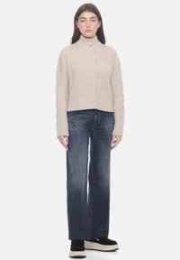 Maglione a coste beige con colletto alto, sei bottoni sul davanti, maniche lunghe e trecce laterali testurizzate, abbinato a jeans blu scuro.