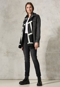 Schwarze und graue Jacke mit strukturiertem Finish, kombiniert mit einem weißen T-Shirt, dunklen Jeans und schwarzen Schnürstiefeln. Der Schal hat ein auffälliges geometrisches Muster.