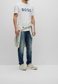 Camiseta blanca con el texto "BOSS" en azul marino, chaqueta de patrón gris claro atada a la cintura, jeans azules y zapatillas blancas con acentos beige.