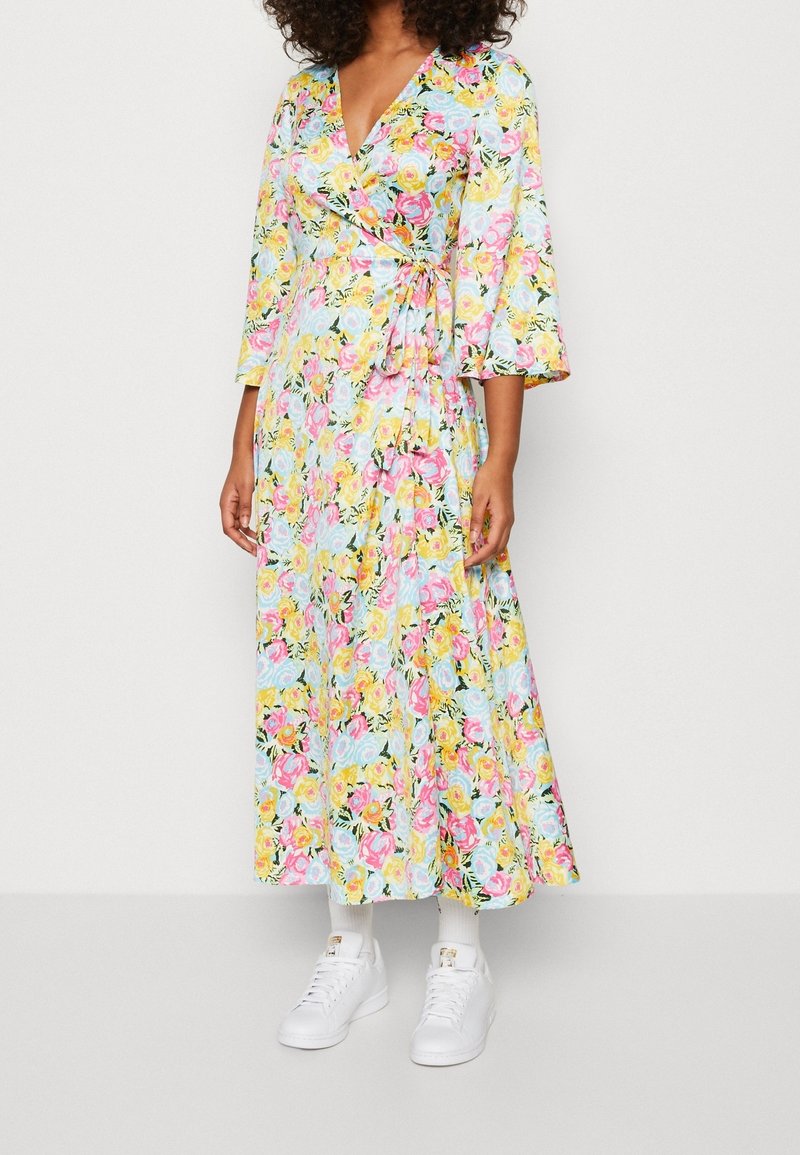 Femme portant une longue robe portefeuille fleurie avec des fleurs roses, jaunes et bleues, associée à des chaussettes blanches et des baskets blanches.