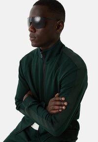 Mand iført en mørkegrøn zip-up jakke og sorte oversized solbriller, med armene over kors, vendende mod venstre på hvid baggrund.