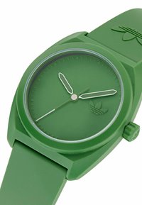 Grön gummiklocka med en slät rund urtavla, minimalistisk design, vita tim- och minutvisare, samt en präglad logotyp på remmen.