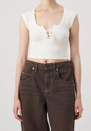 Kvinde iført hvid cropped top med snøring og højtaljede mørkebrune jeans, stående mod en ensfarvet baggrund.