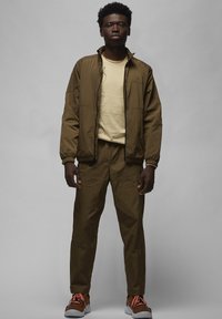 Brun bomberjakke med elastiske manchetter, beige grafisk skjorte, matchende bukser og brune sneakers med orange accenter. Glat tekstur, afslappet pasform.