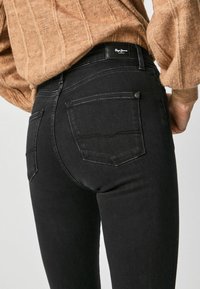 Jeans de mezclilla negra con un diseño ajustado, que cuentan con dos bolsillos traseros, detalles de costura sutiles y una etiqueta de marca en la cinturilla.
