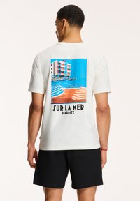 Balta medvilninė marškinėlių su išskirtiniu spalvingu paplūdimio peizažu ant nugaros, su tekstu "SUR LA MER BIARRITZ" drąsiomis juodomis raidėmis.