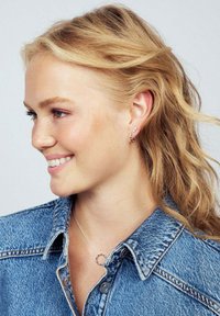 Silverfärgade hoopörhängen och ett delikat silverhalsband med en cirkulär hängande berlock, använt över en jeansjacka. Mjuka vågiga hårslockar ramar in ansiktet.