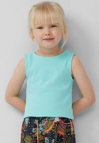 Jong kind met blond haar, gekleed in een lichtblauwe mouwloze top en een gestreepte rok, poseert met de handen achter de rug tegen een grijze achtergrond.