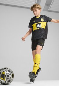 Camiseta de fútbol negra y amarilla con logo, pantalones cortos negros y calcetines amarillos. Jugador en acción, pateando un balón de fútbol con patrón.