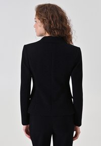 Blazer noir sur mesure avec texture en chevron, épaules structurées et silhouette ajustée. Comprend un détail discret de basque à la taille.