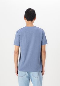 Μπλε κοντομάνικο πουκάμισο με υφασμάτινο σχέδιο πλέγματος, στρογγυλή λαιμόκοψη και πλαϊνές σχισμές, σε συνδυασμό με ανοιχτό μπλε τζιν παντελόνι.