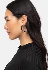 Boucles d'oreilles en acrylique noir et transparent avec un motif marbré, de forme circulaire et finition brillante, portées dans une vue de profil.