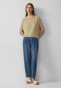 Beige T-shirt met korte mouwen en een wijde halslijn, gecombineerd met losse blauwe denim jeans en witte sandalen met een gespsluiting. Eenvoudige en relaxte pasvorm.