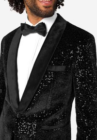 Moški se smeji, oblečen v črno bleščečo smoking jakno, belo nagubano srajco in črno metuljčkasto kravato na beli podlagi.