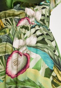 Gros plan d’un tissu vert avec une couture élastique et un imprimé floral coloré mettant en valeur de grandes feuilles et des fleurs bordées de rose.