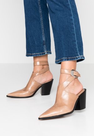 Ben i cropped blå jeans iført beige læder sandaler med spidse tæer, blokhæl, ankelremme og sølvspænder på hvidt gulv.