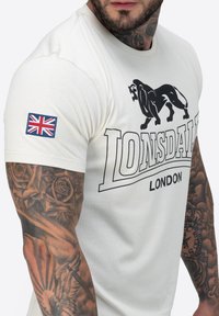 Majica krem boje s uskim krojem, s crnim grafičkim otiskom lava i natpisom "Lonsdale London". Sadrži oznaku britanske zastave na rukavu.