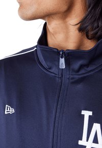 Marineblaues Zip-Up-Jackett mit hohem Kragen, dezenten weißen Streifen und weißem gesticktem Logo auf der Vorderseite. Glatte, sportliche Stofftextur.