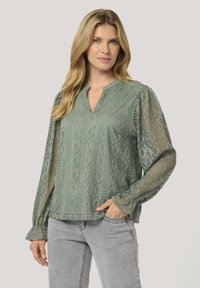 Blusa verde chiaro con scollo a V, maniche trasparenti a motivo, design a diamante testurizzato e polsini arricciati. Indossata con jeans grigi chiari.