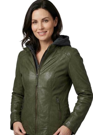 Frau mit dunklen Haaren lächelnd, trägt eine olivgrüne Lederjacke mit Frontreißverschluss und eine dunkle Kapuzenlage darunter.