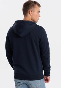 Námořnická mikina s texturovanou látkou; má stahovací kapuci, žebrované manžety a pas. Pohled zobrazuje zadní design a volný střih.