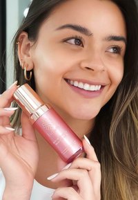 Bottiglia di siero rosa con accenti metallici dorati, forma curvata e finitura texturizzata, tenuta da una persona con unghie lunghe e orecchini.