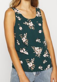 Femme portant un débardeur vert foncé à imprimé floral rose et blanc, associé à un jean en denim bleu clair.