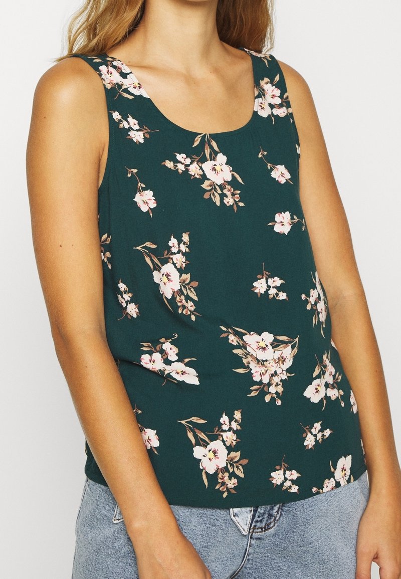 Femme portant un débardeur vert foncé à imprimé floral rose et blanc, associé à un jean en denim bleu clair.