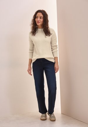 Pull côtelé crème avec un col montant, associé à un jean droit bleu foncé et des baskets beiges sur un fond neutre.