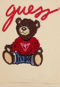 Σχέδιο αρκούδας με κεντημένα σχέδια σε κρέμ φόντο, φορώντας κόκκινη μπλούζα με τριγωνικό λογότυπο και μπλε σορτς, με υφή που αναδεικνύει τις λεπτομέρειες της γούνας.