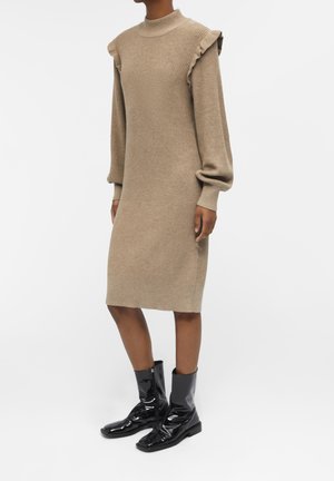 Kvinde iført en beige ribstrikket midikjole med flæsedetaljer på skuldrene og sorte skinnende midtkalvstøvler står mod en hvid baggrund.