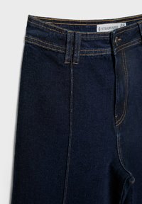 Tmavě modré džíny z denimu s hladkou texturou, s kontrastním šitím, zapínáním na knoflík a středním pasem. Viditelný štítek velikosti 38.