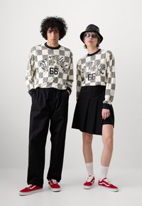 Camicie a maniche lunghe a scacchi in crema e nero con grafiche, abbinate a pantaloni neri e a una gonna. Entrambi indossano sneaker rosse.