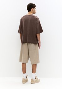 T-shirt oversize marrone, pantaloni corti beige, tasca posteriore visibile, calzini bianchi, sneakers beige chiaro. Lo sfondo semplice mette in risalto l'outfit.