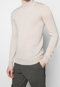 Béžový rolák na osobě s jednou rukou v kapse tmavě šedých kalhot na jednobarevném světlém pozadí.