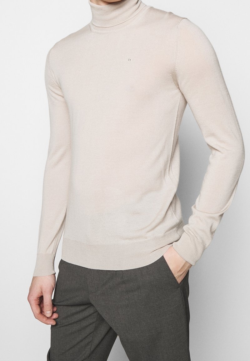 Béžový rolák na osobě s jednou rukou v kapse tmavě šedých kalhot na jednobarevném světlém pozadí.