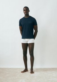 Meztelen lábú férfi áll egy szőnyegen, sötétkék pólót és fehér boxeralsót visel egyszínű világosszürke háttér előtt.