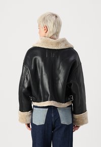 Veste en cuir noir avec un col et des poignets en shearling beige pelucheux, présentant un design court et des accents à boucle à la taille.