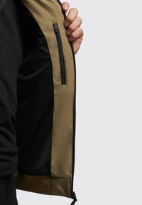 Khaki jas binnenkant met een zwarte mesh voering en een ritszak, met een gladde textuur en contrasterende kleuren.